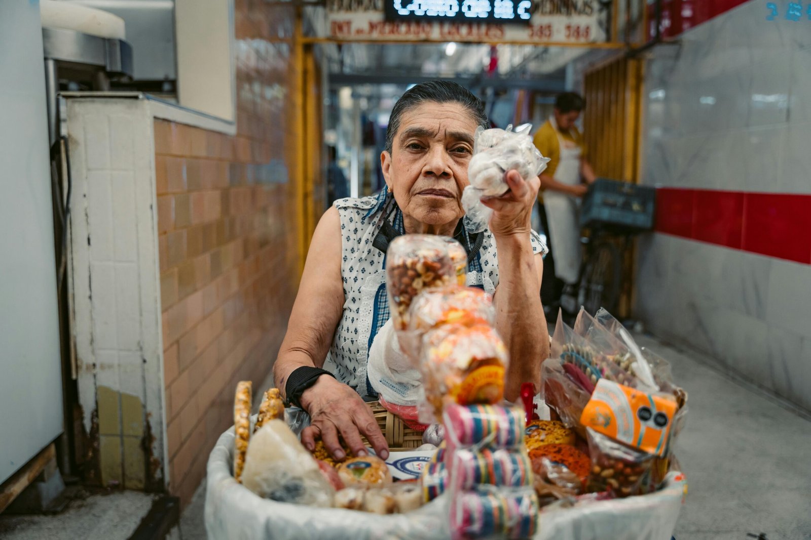 A senior vendor in a market alley in Mexico City displaying colorful street food products.