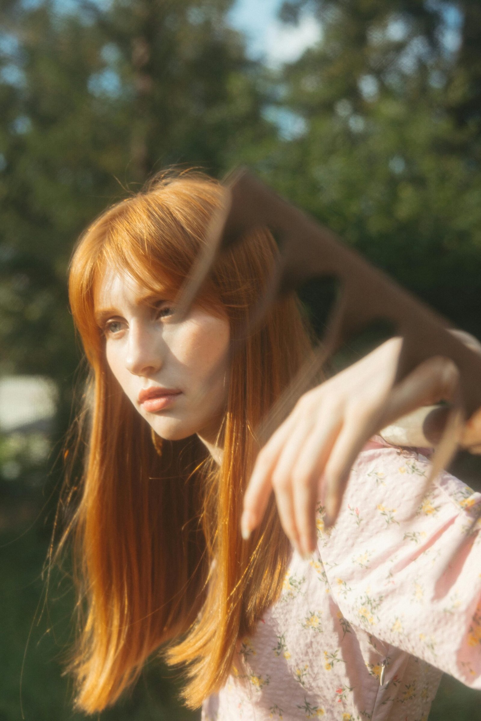 Artistic portrait of a red-haired woman in a floral dress with a blurry hand in the foreground.