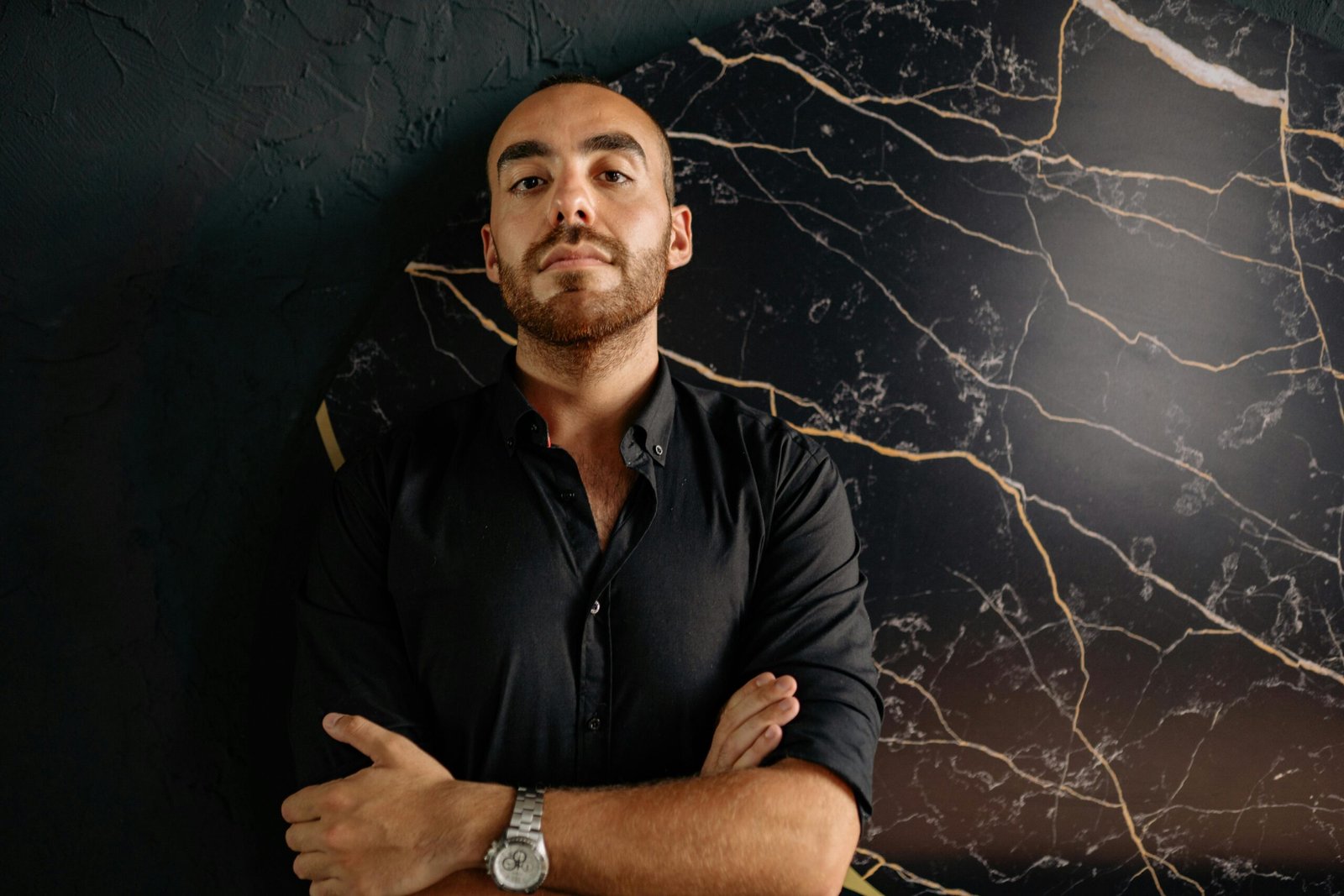 Portrait of a confident man with crossed arms in black attire against a marble background.