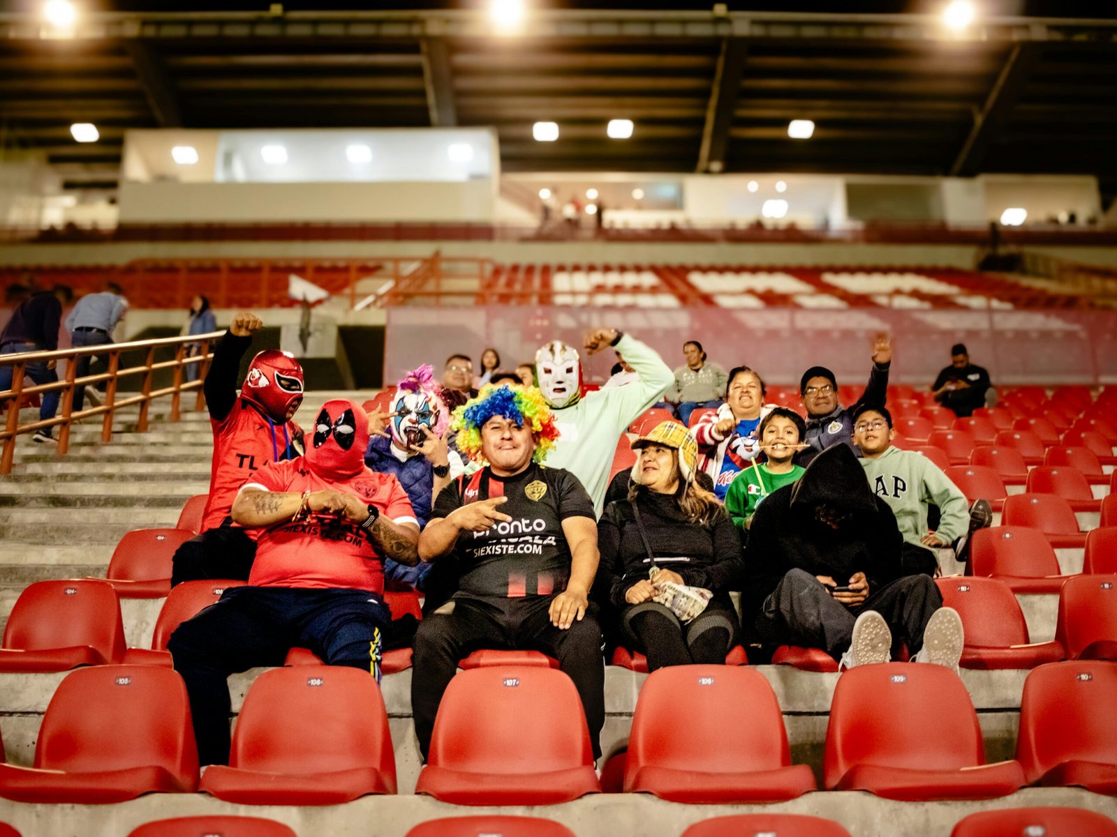 Group of colorful football fans cheering in stadium bleachers at night.